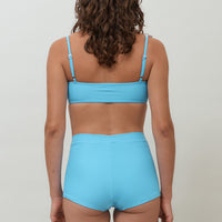 Woman wearing blue high waist surf shorts, sustainable swimwear designed to stay on in the surf, offering supportive coverage and a flattering fit for active beach days.