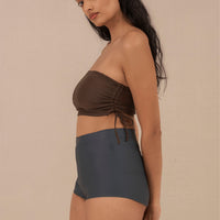 Woman wearing blueish grey high waist surf shorts, sustainable swimwear designed to stay on in the surf, offering supportive coverage and a flattering fit for active beach days.