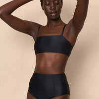 A high-waist black sustainable swimwear bikini bottom with minimalist design and moderate coverage, modelled by a woman in a matching bikini top.