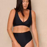 A high-waist black sustainable swimwear bikini bottom with minimalist design and moderate coverage, modelled by a woman in a matching bikini top.